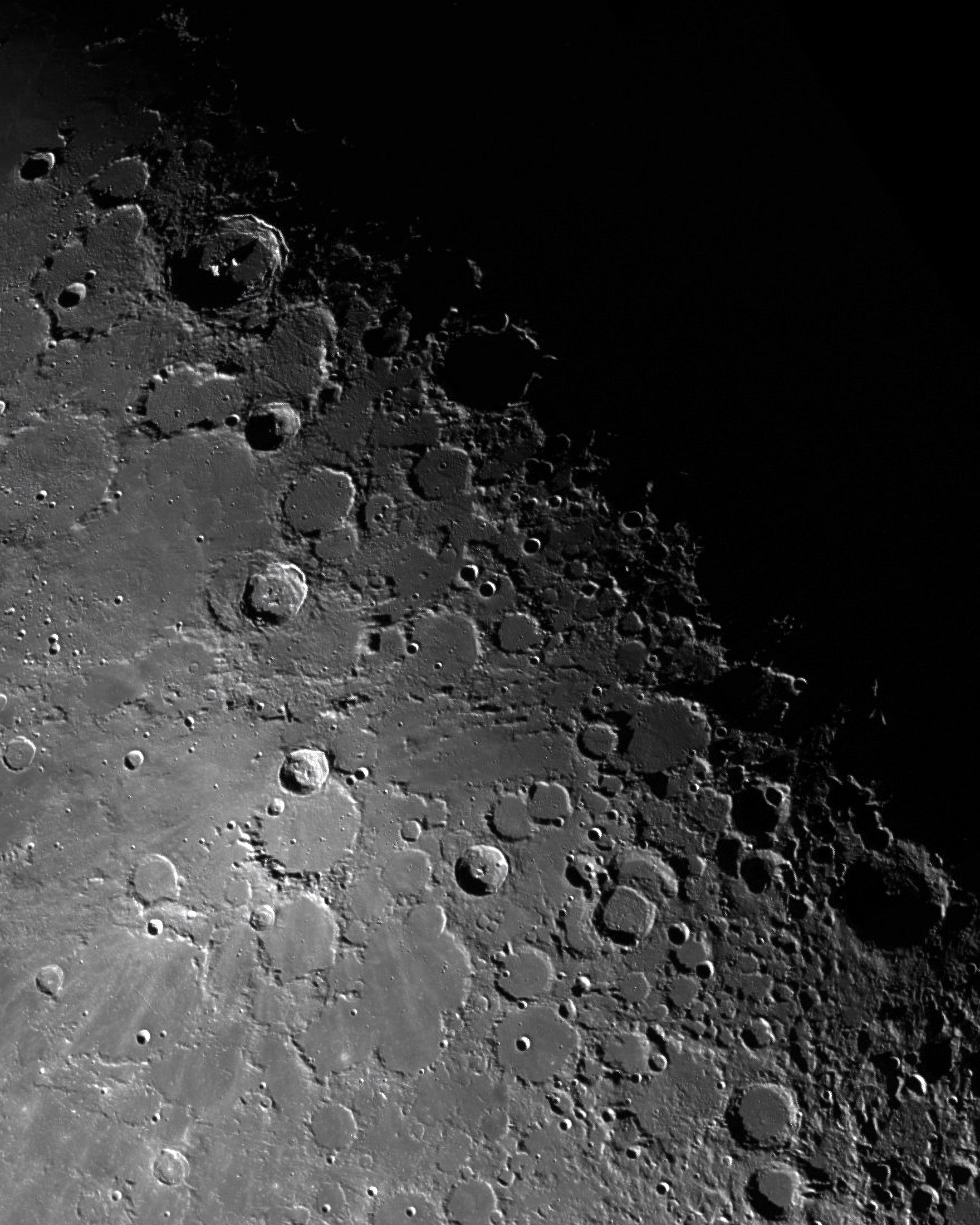 A black-and-white close-up of the cratered moon surface with many impact basins and a large black void in the upper right.
