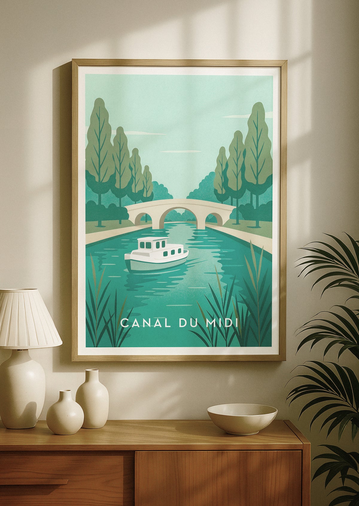 Plakat podróżniczy w ramie przedstawiający Canal du Midi we Francji, z retro łodzią na turkusowej wodzie, drzewami i kamiennym mostem. Wisi na beżowej ścianie nad drewnianą konsolą z ozdobnymi wazonami i lampą.